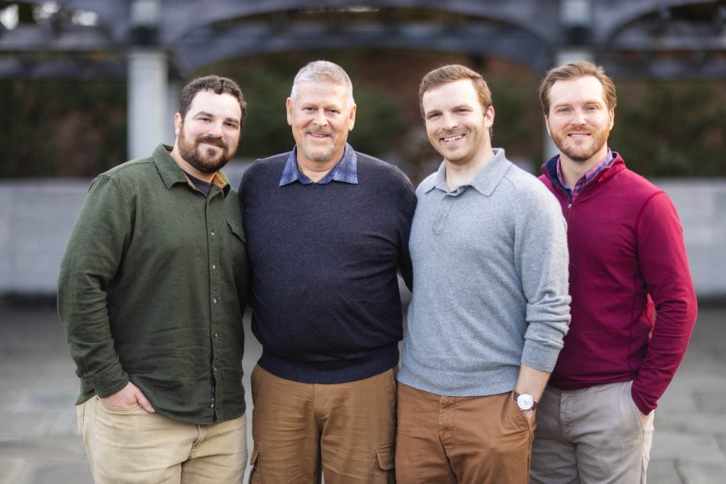 Four men standing outdoors, smiling at the camera, with the Jefferson Memorial in the background. The man on the left wears a green shirt, the second man wears a blue shirt, the third man wears a gray shirt, and the fourth man wears a red shirt. It's as if they planned their own family portrait session.