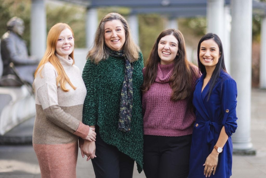 Four women standing outdoors, smiling at the camera. They are dressed in casual yet stylish outfits and are standing in front of a statue and pillars at the Jefferson Memorial, creating a charming family portrait.