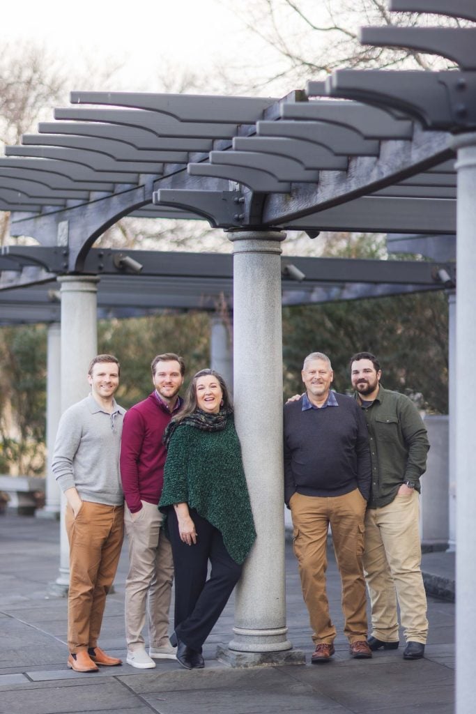 Five adults stand under a pergola structure outdoors on a paved area, smiling for a group photo. Four men and one woman are casually dressed and evenly spaced around the columns, creating a charming family portrait reminiscent of classic scenes at the Jefferson Memorial.