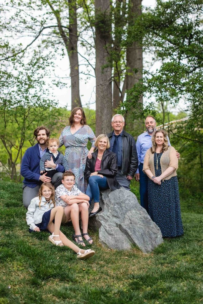 An extended family of nine, including adults and children, pose together outdoors on a grassy area at Brookside Gardens, surrounded by trees and rocks.