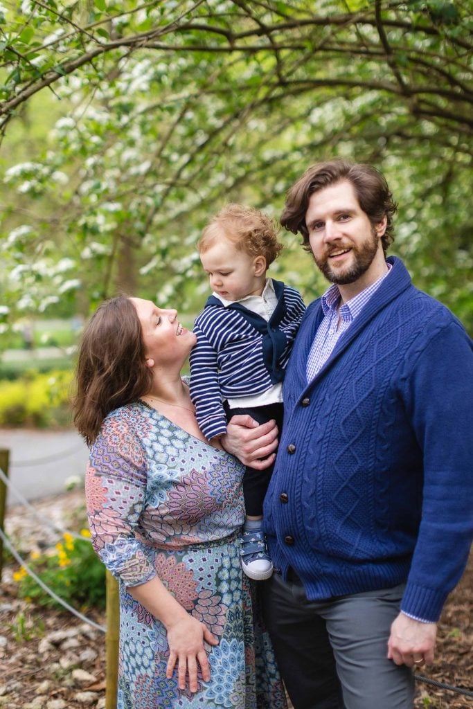 A family of three poses for a portrait under a tree. The man holds a baby as the woman looks up at the child, embodying joy. They are all dressed in bluetoned clothing, capturing an extended moment of connection and warmth.