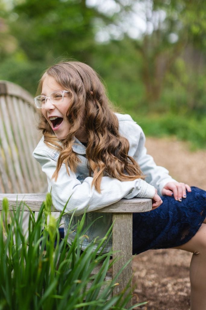 A young girl with long wavy hair and glasses, wearing a denim jacket, sits on a wooden bench outdoors, smiling widely for a portrait that captures the essence of joy in her extended family.