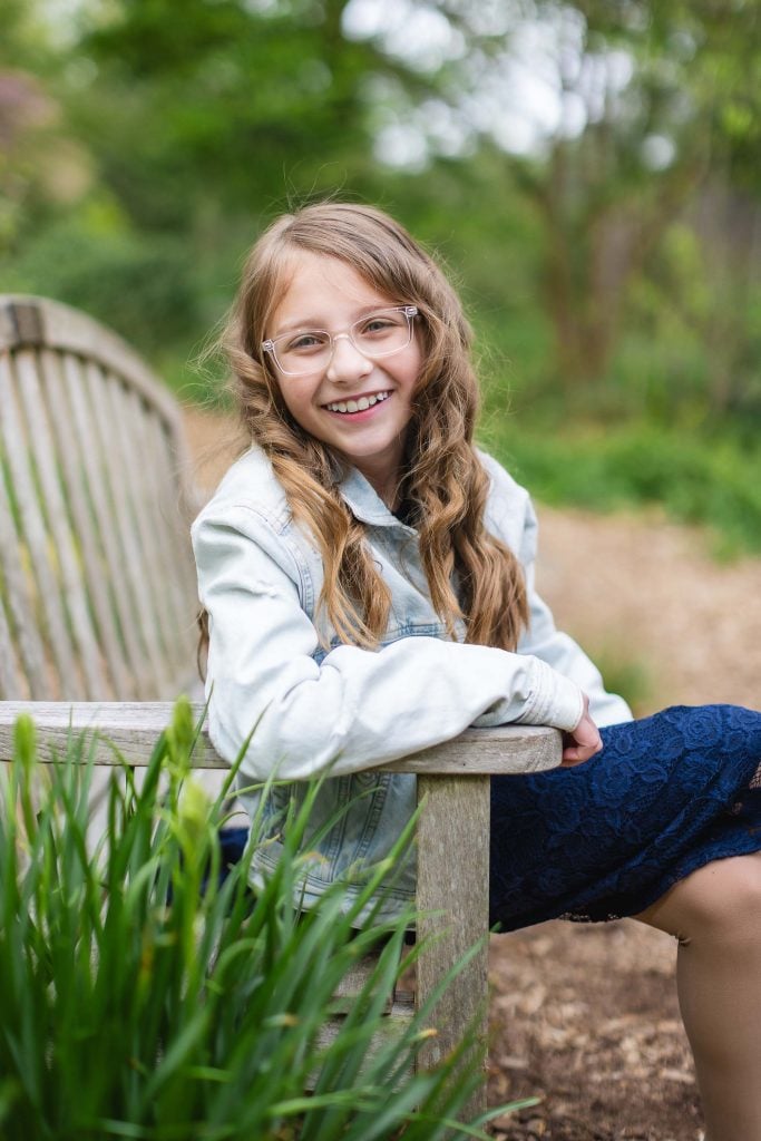 A young girl with long blonde hair and glasses sits on a wooden bench outdoors, smiling and leaning on the bench arm. This charming portrait features greenery and a garden path in the background, capturing a warm family moment.