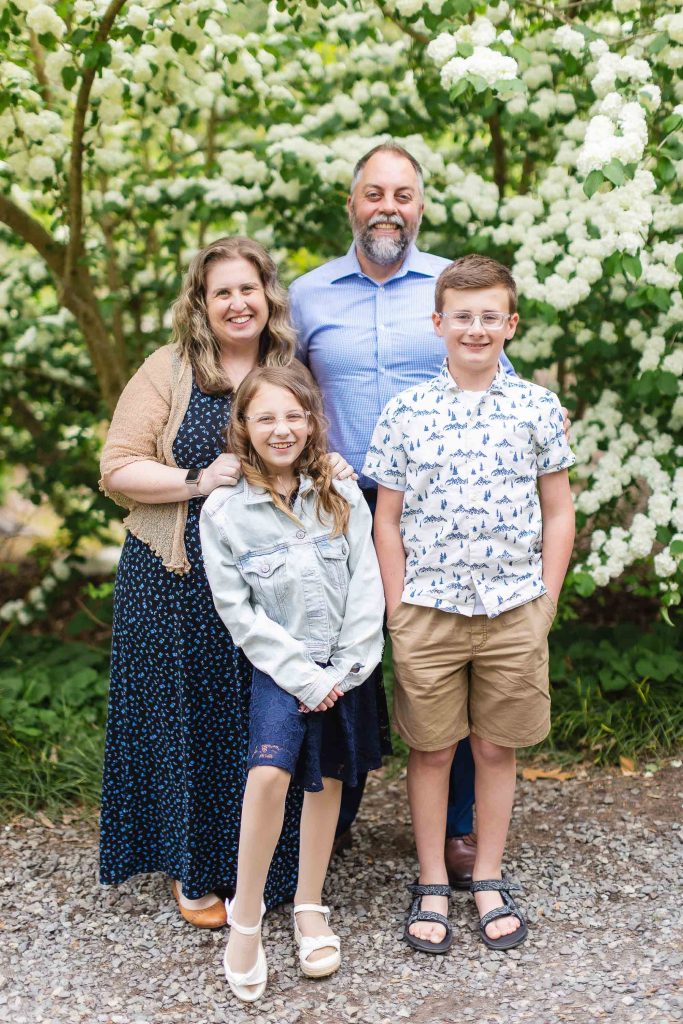 An extended family of four standing outdoors in front of a flowering bush. The parents stand behind their two children, all four are smiling at the camera, creating a heartwarming portrait.