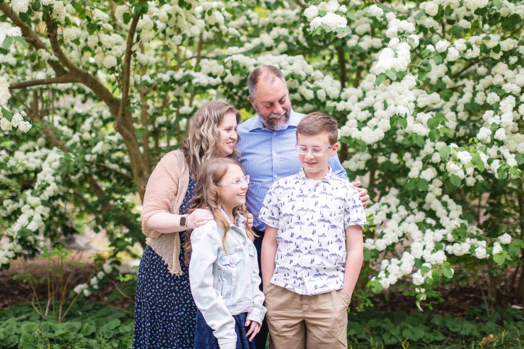 A family of four, including two adults and two children, stands together in front of white flowering bushes. They are smiling, the children are wearing glasses, and the entire portrait captures a moment of pure joy.