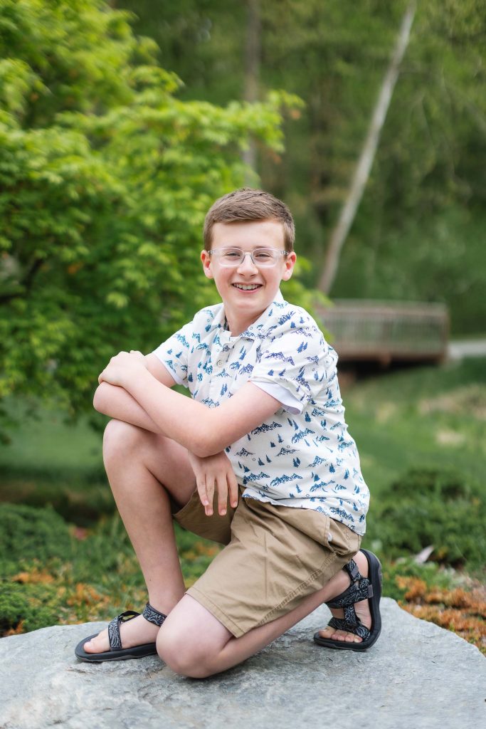 A young boy wearing glasses, a patterned shirt, khaki shorts, and sandals is kneeling on a rock with greenery in the background at Brookside Gardens.