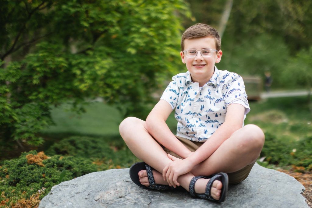 A portrait of a boy with glasses sitting crosslegged on a rock, wearing a white shirt with blue patterns and shorts, outdoors at Brookside Gardens, framed by the lush trees in the background.