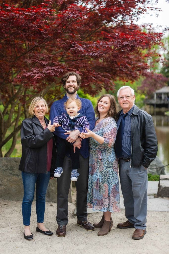 An extended family of five, including an elderly couple, a man holding a baby, and a woman, pose outdoors in front of a tree with red leaves for a heartwarming portrait.