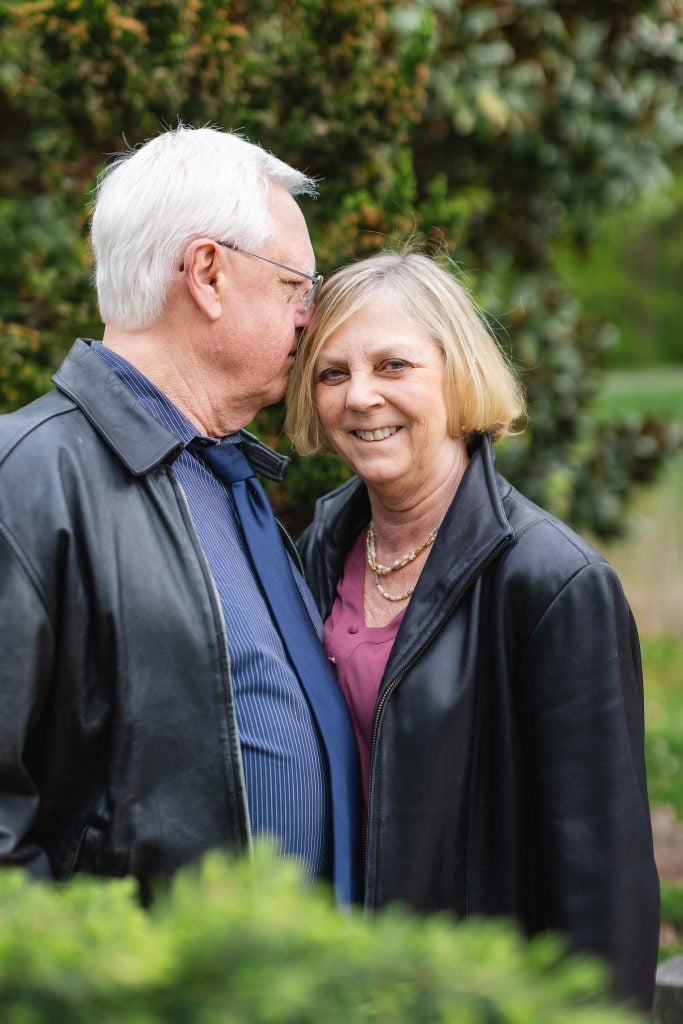 An elderly man with white hair whispers to a smiling elderly woman with blonde hair. They stand outdoors at Brookside Gardens, both wearing black jackets. The extended greenery in the background adds a refreshing touch to this charming portrait.