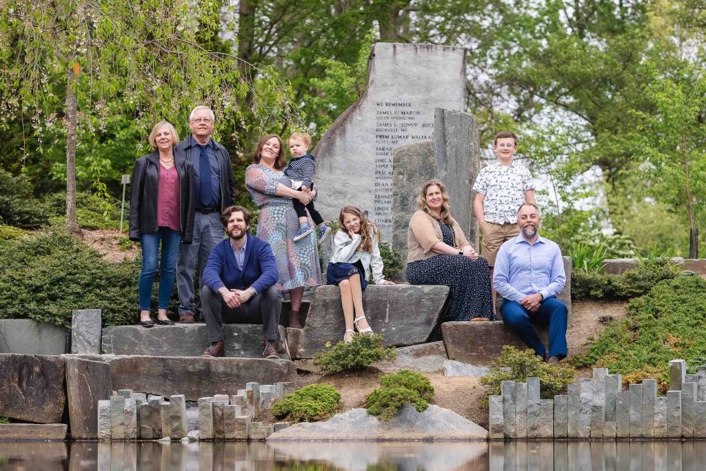 An extended family of eight poses outdoors in front of a stone monument, surrounded by trees and greenery, creating a charming portrait.