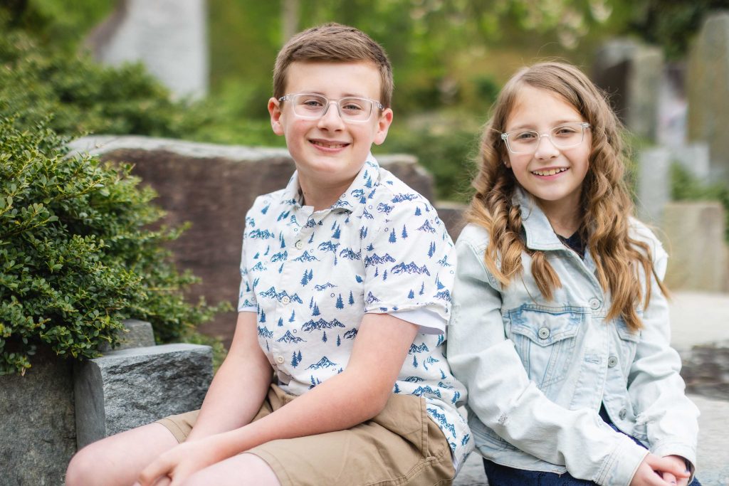 Two children with glasses, one boy and one girl, are sitting outdoors on a stone bench, smiling at the camera in this charming family portrait. The boy is wearing a white shirt with a mountain print and shorts.