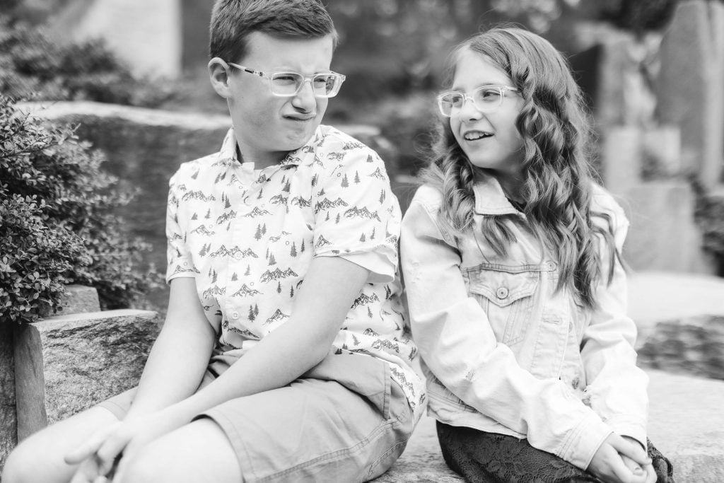 A black and white portrait of two children sitting on a bench at Brookside Gardens. The boy on the left wears glasses and makes a funny face, while the girl on the right, also wearing glasses, looks at him and smiles.