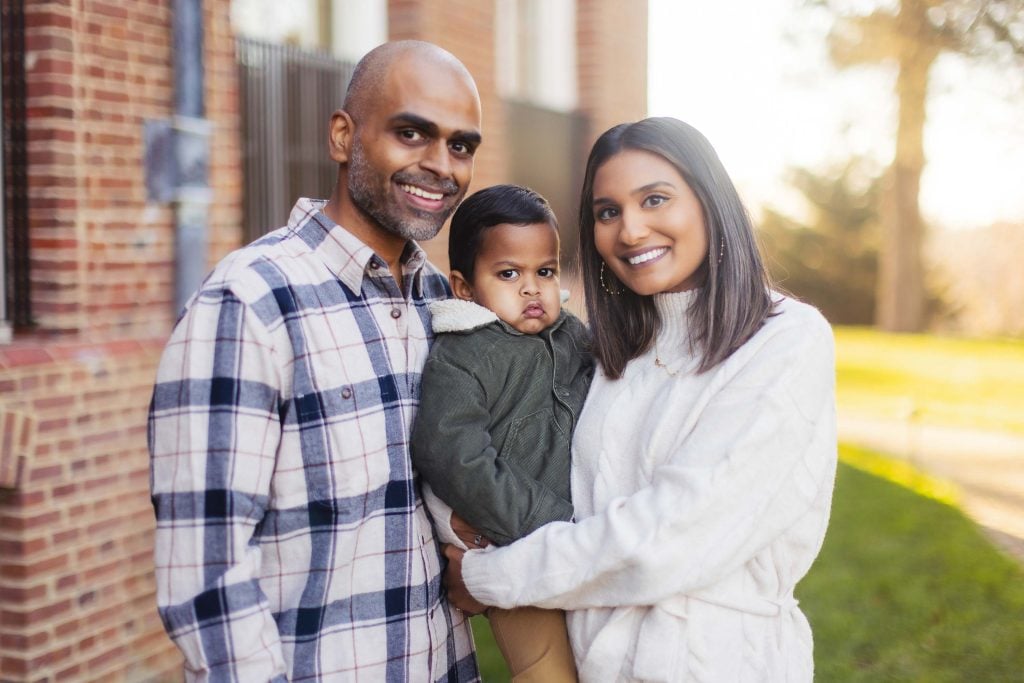 A smiling family stands outdoors holding a young child. The man wears a plaid shirt, the woman a white sweater, and the child a green jacket. Brick building and greenery are in the background, capturing an idyllic moment at Belair Mansion.