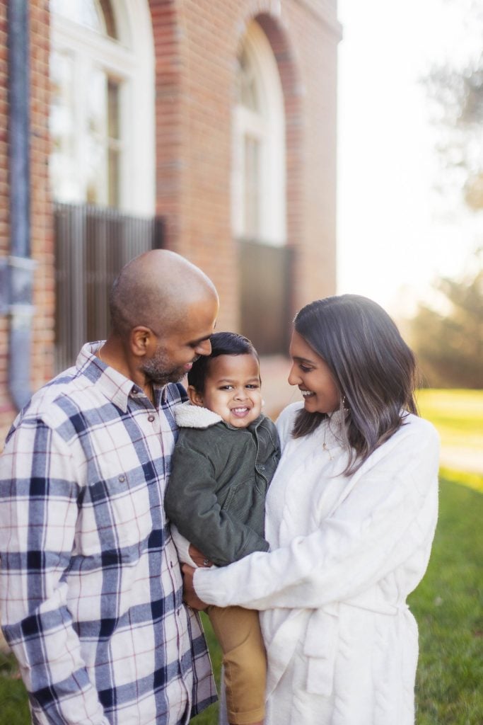 A family of three stands outdoors near a brick building. The man holds a young boy who smiles at the camera, while the woman, also smiling, looks at the boy. All are dressed warmly in this charming portrait that captures their joy and togetherness.