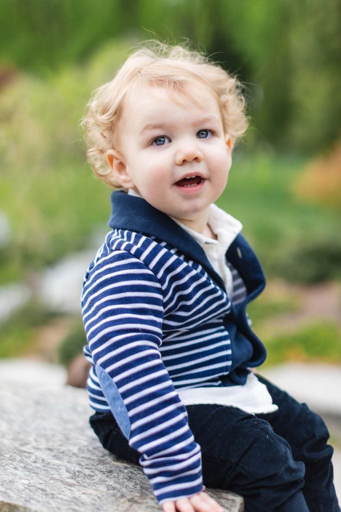 A toddler with curly blond hair, wearing a blue and white striped sweater, sits outdoors on a stone surface at Brookside Gardens with a blurred green background.