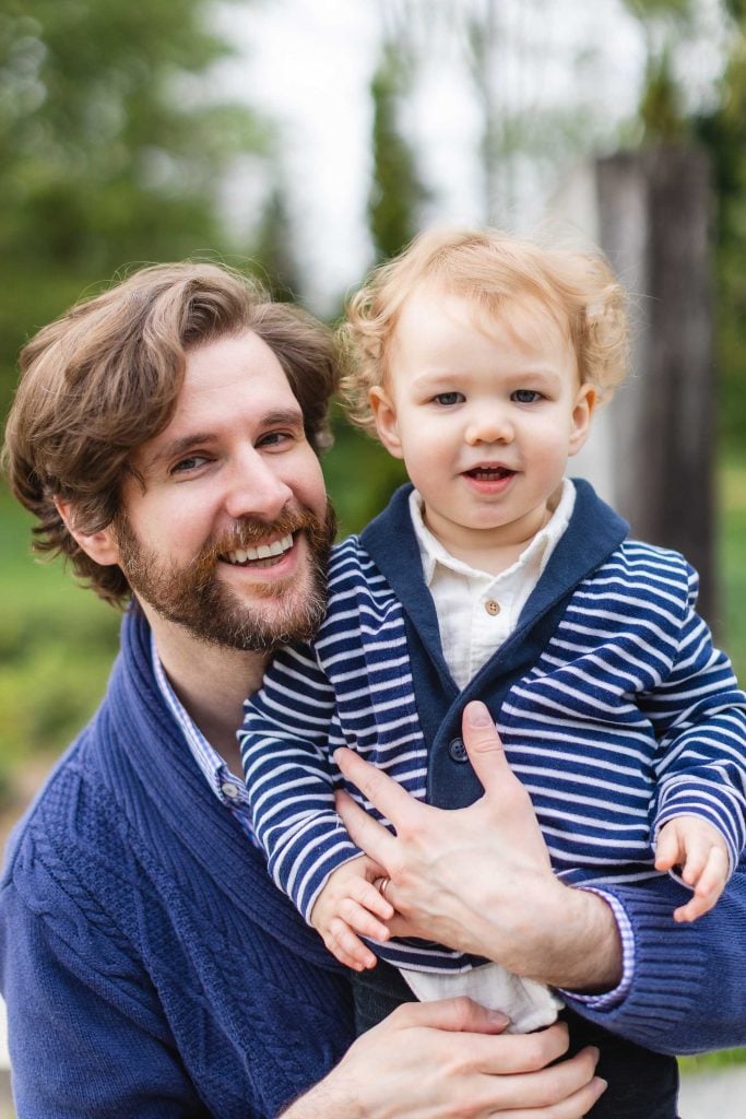 In this heartwarming portrait at Brookside Gardens, a bearded man holds a young child wearing a striped sweater. They are both smiling, surrounded by lush greenery in the background.