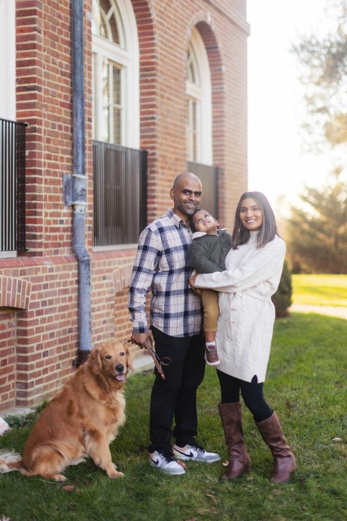 A family stands outside the Belair Mansion, framed by its brick façade and arched windows. The parents smile, holding their toddler, as a golden retriever sits beside them on the manicured grassy lawn—a perfect portrait of love and togetherness.