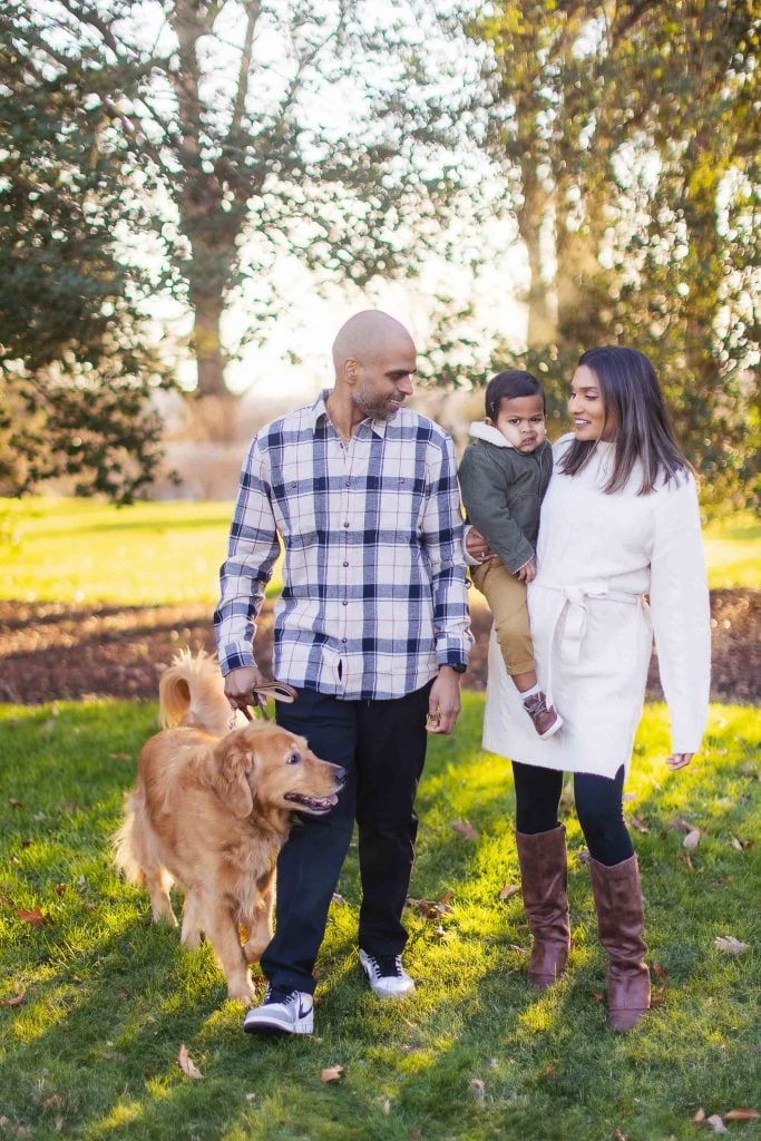 A family of three, with a man and a woman holding their child, accompanied by their dog, walking together in Belair Mansion's park on a sunny day.