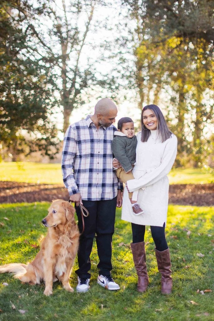 A family with a toddler and a dog stands outdoors. The man is holding the child's hand, the woman is holding the child, and the dog is sitting beside them. It is a sunny day with trees in the background, making for perfect outdoor family portraits.