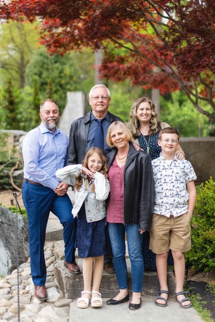 A group portrait of six people, including three generations, taken outdoors at Brookside Gardens near trees and rocks. Two adults stand in the back row, while two adults and two children stand in the front row.