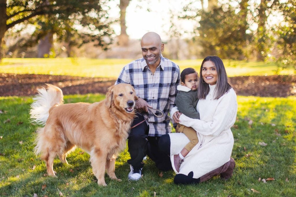 A family poses outdoors with a golden retriever, capturing a beautiful family portrait. The father holds a leash and smiles, while the mother kneels beside him holding their young child. Trees and sunlight provide a stunning backdrop, reminiscent of charming moments at Belair Mansion.