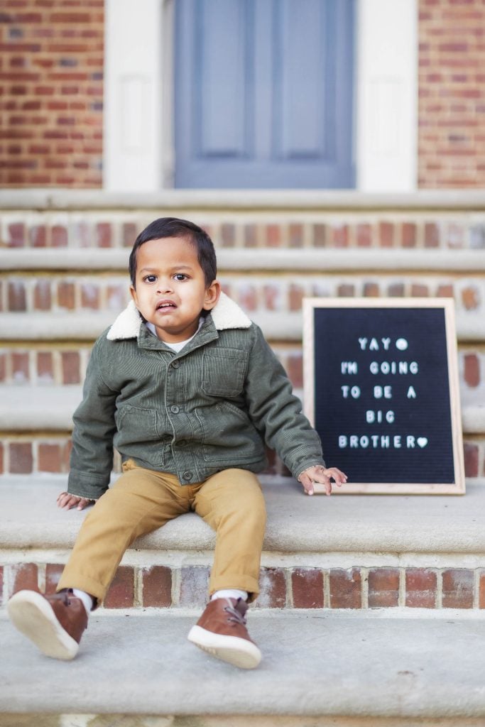 A young child sits on the steps of Belair Mansion with a sign next to them that reads, "Yay, I'm going to be a big brother." The child is dressed in a green jacket and beige pants, capturing a heartwarming moment perfect for maternity portraits.