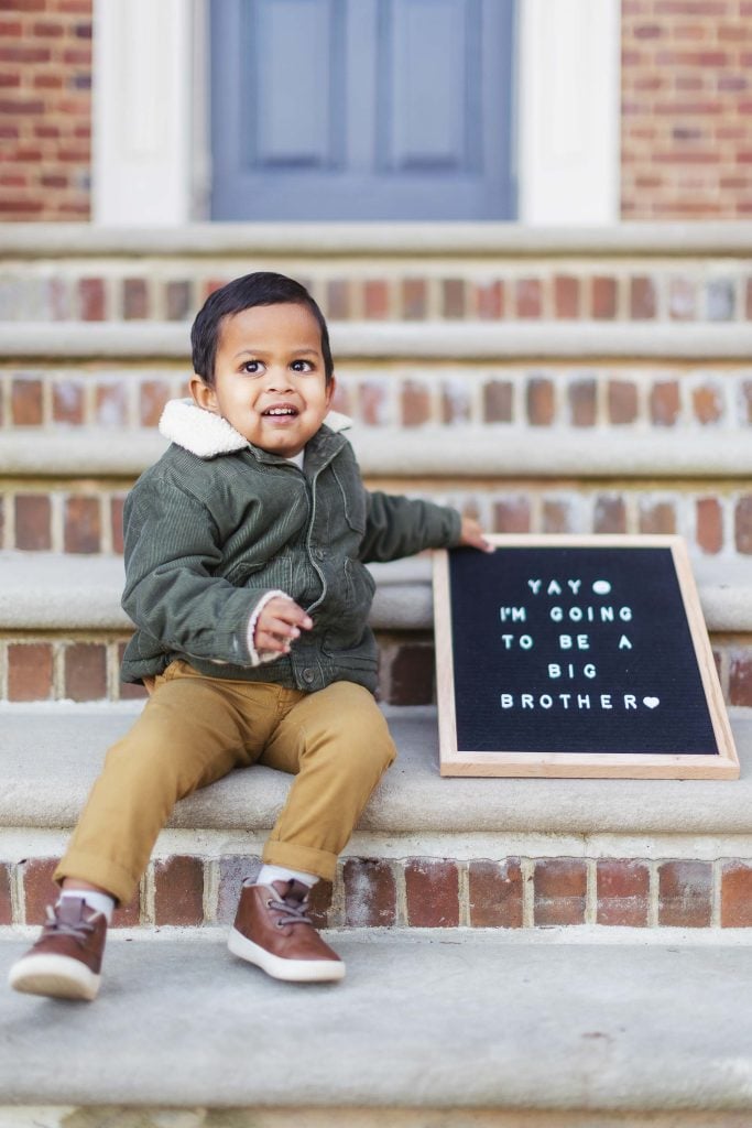 A young child sits on steps holding a sign that reads, "Yay, I'm going to be a big brother." The family portrait shows the child in a green jacket, tan pants, and brown shoes, with a blue door and brick wall of Belair Mansion in the background.