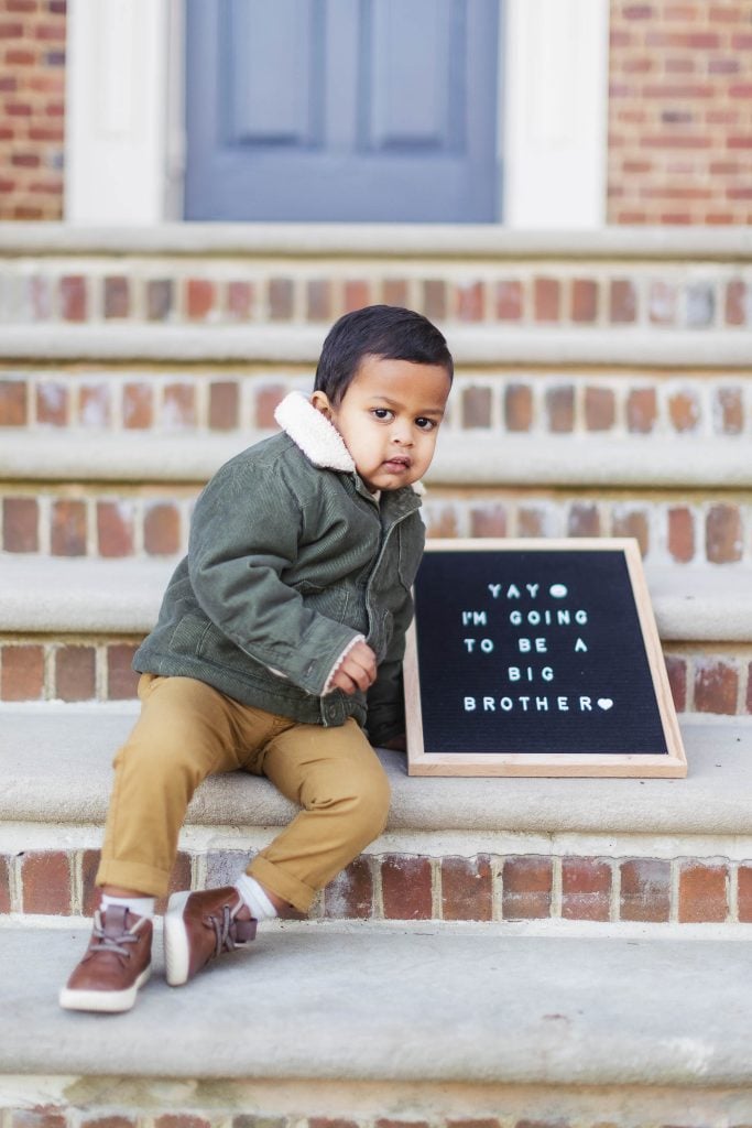 A young child sits on brick steps at Belair Mansion next to a letter board displaying the message, "YAY ♥ I'M GOING TO BE A BIG BROTHER ♥." This heartwarming portrait captures the joy and anticipation of maternity news.