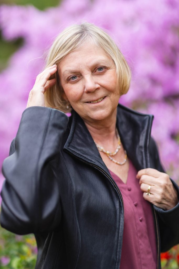 A woman with short blonde hair wearing a black jacket and maroon top stands in front of the vibrant pink flowers at Brookside Gardens, smiling and holding her hair with one hand.