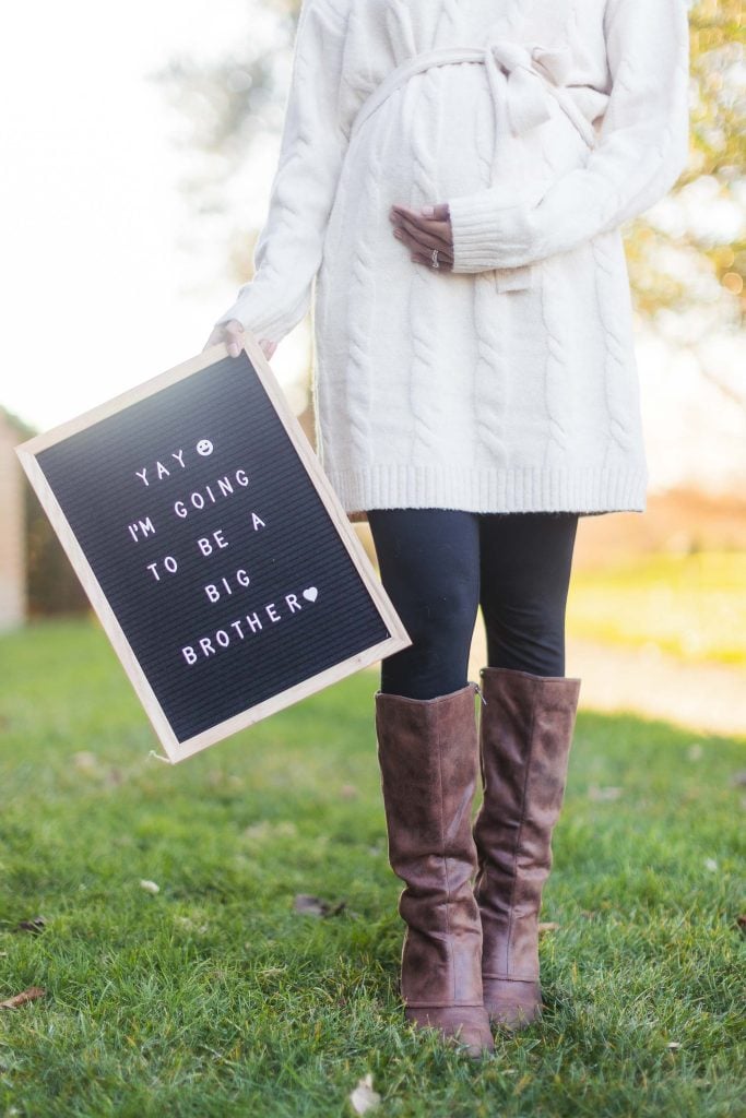 A person wearing a white sweater and brown boots holds a letter board sign that reads, "Yay, I'm going to be a big brother," while standing on grass at the Belair Mansion. Their hand is lovingly resting on their pregnant belly, celebrating this special family moment.