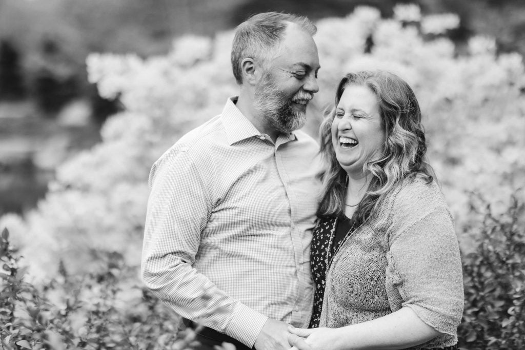 A man and a woman, standing outside amidst foliage at Brookside Gardens, are smiling and laughing while holding hands. The image is in black and white.