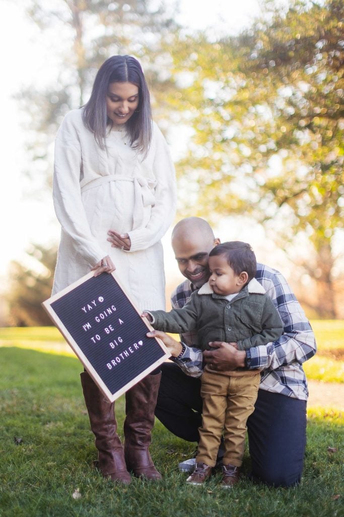 In a grassy outdoor setting at Belair Mansion, a woman holds her pregnant belly while standing next to a kneeling man and young boy. The boy points to a letter board that reads, "Yay, I'm going to be a big brother," capturing the joyous moment in their family portrait.