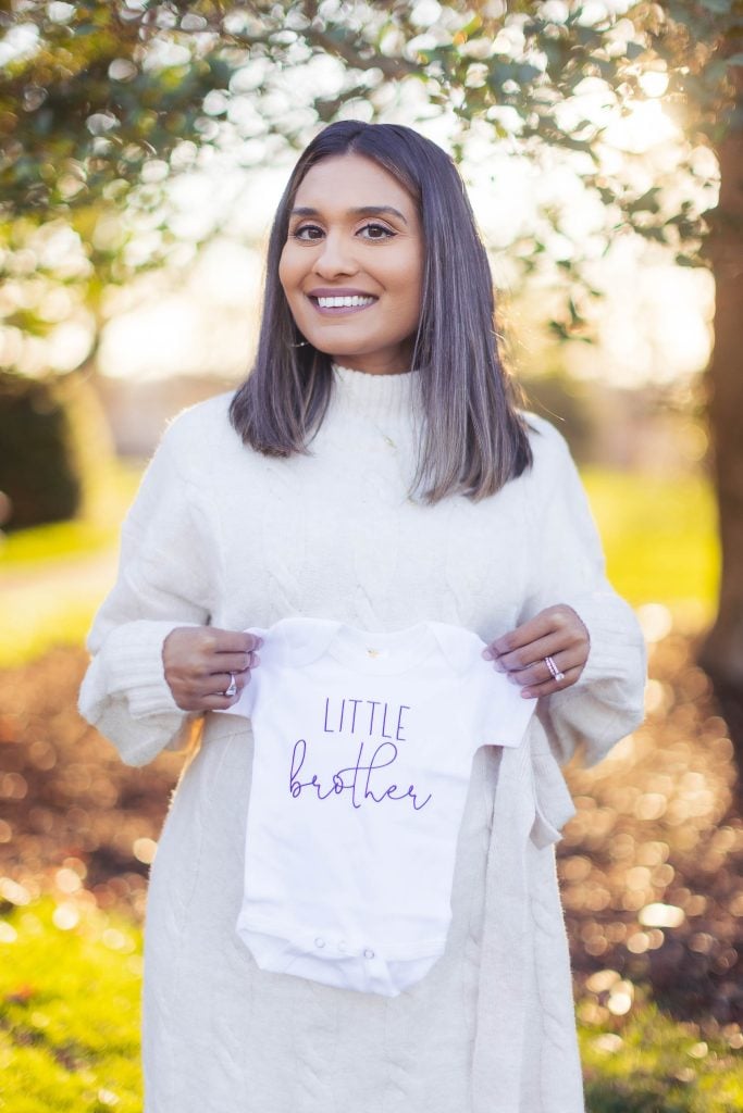 A woman is standing outside Belair Mansion, holding a baby onesie that says "Little brother." She is smiling and wearing a white sweater, capturing a perfect family portrait.