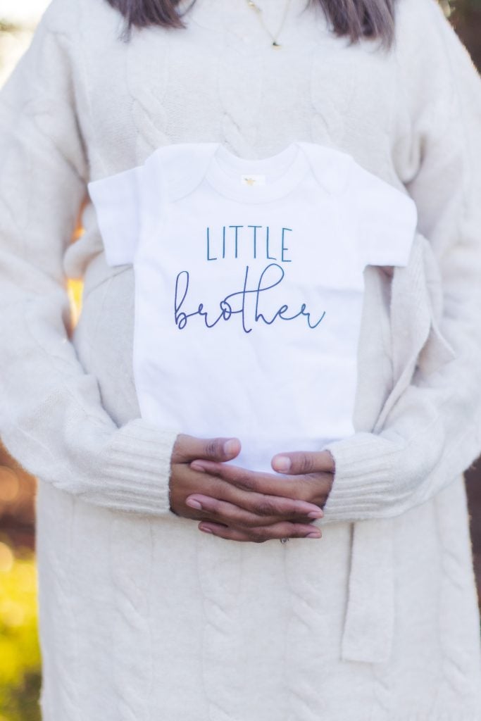 A person wearing a white sweater stands in the elegant Belair Mansion, holding a small white onesie with the text "Little brother" written on it, celebrating a joyous moment of family and maternity.