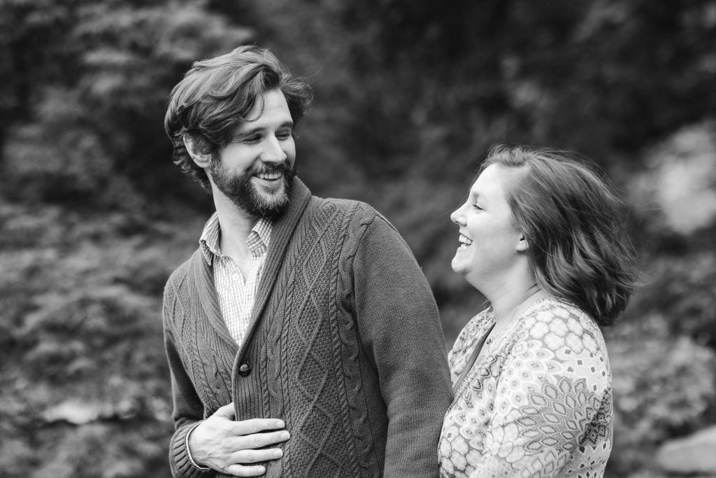 Black and white image of a man and a woman outdoors at Brookside Gardens, engaged in conversation and smiling. The man is wearing a cardigan and the woman is in a patterned sweater. Trees are blurred in the background, giving it an intimate, family gathering vibe.