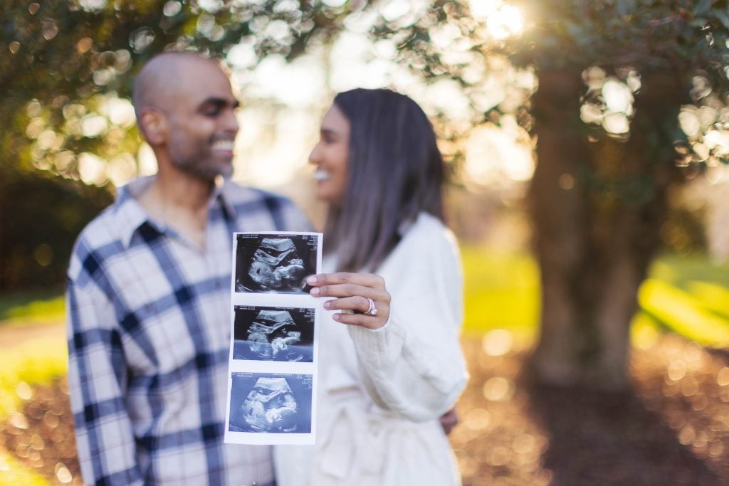 A smiling couple holds up ultrasound images outdoors, indicating a maternity moment at Belair Mansion. The background is blurred with greenery and sunlight, radiating warmth and joy.