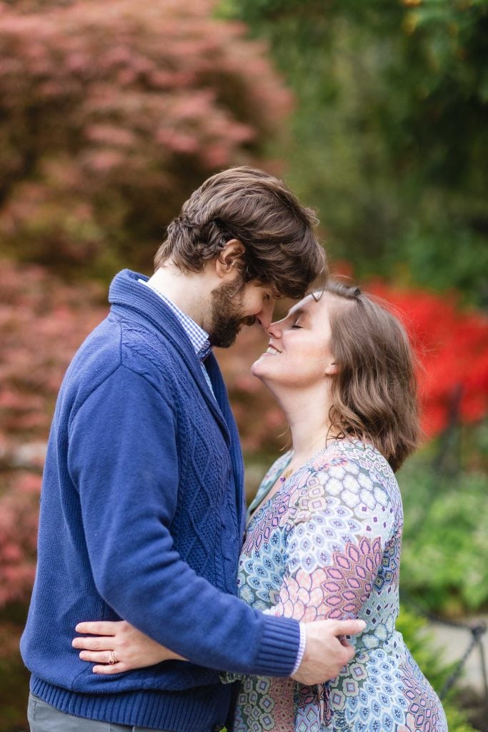 A couple standing close, smiling with their noses touching. They are outside, surrounded by greenery and trees with autumn foliage. Both look happy and are embracing each other in this intimate portrait, capturing a treasured family moment.