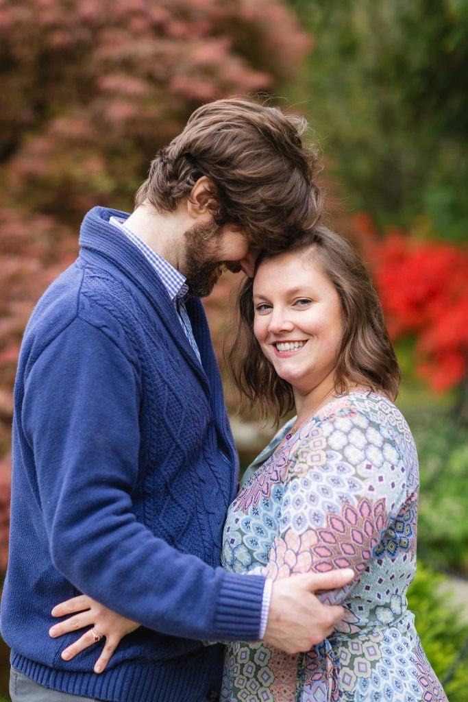 A woman smiles at the camera while a man, eyes closed, leans his head against hers. They are standing outdoors with greenery and red flowers in the background, creating a picturesque portrait of extended family moments.