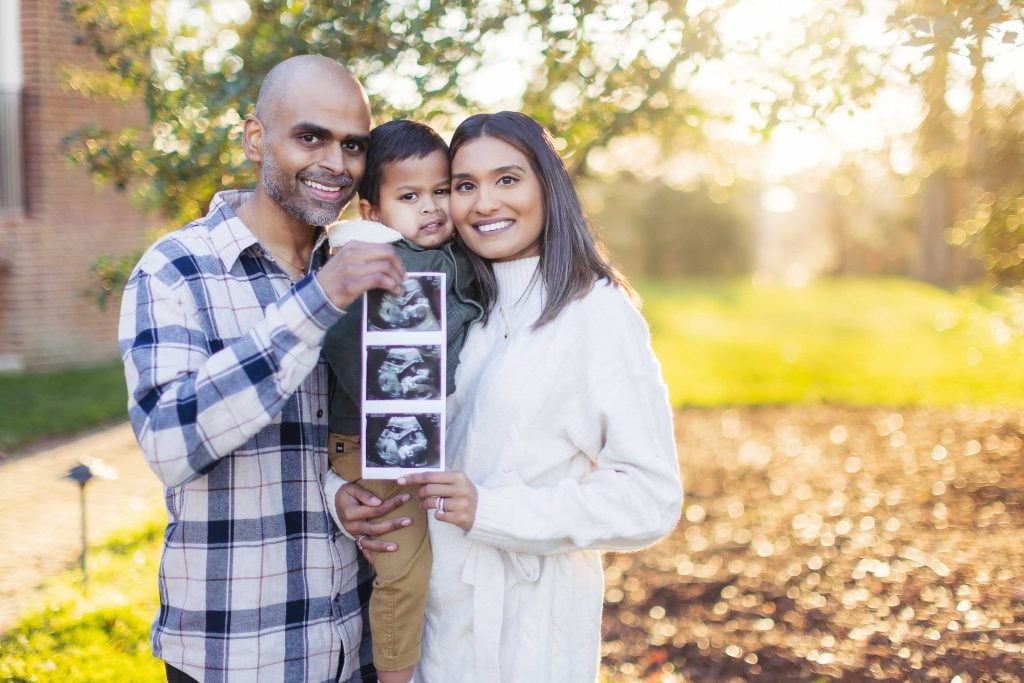 A smiling couple holds their young child and a series of ultrasound images, standing outdoors on a sunny day, perfect for family portraits at Belair Mansion.