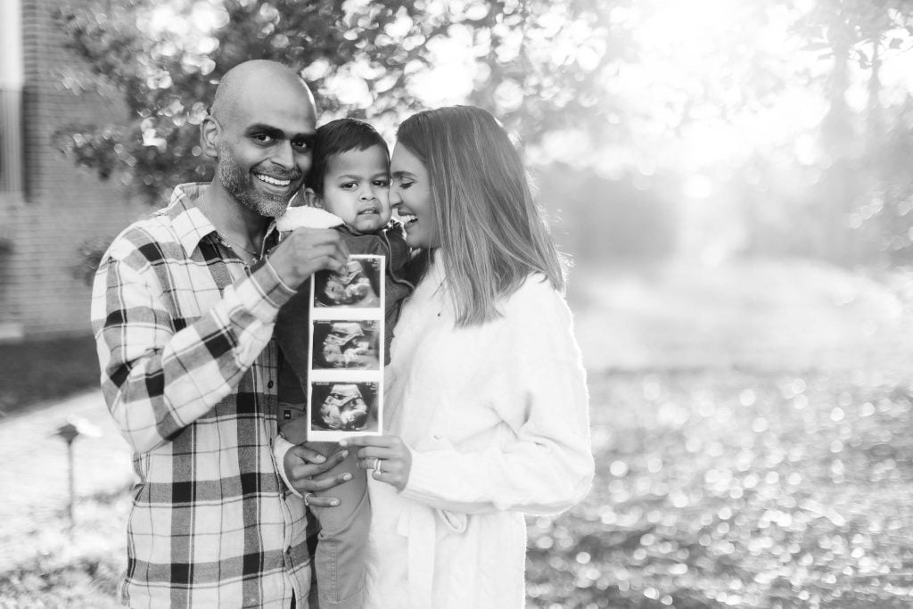 A family stands together outdoors, smiling. The man holds an ultrasound image, hinting at their joyous maternity journey. Bright sunlight filters through the trees in the background of Belair Mansion. The image is in black and white.