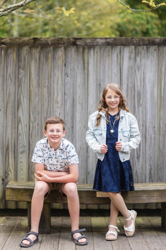 Two children pose outdoors in this charming portrait. One boy sits on a bench, arms resting on his knees, while a girl stands beside him, wearing a denim jacket over a dress. The serene setting of Brookside Gardens and the wooden fence in the background complete this lovely family moment.