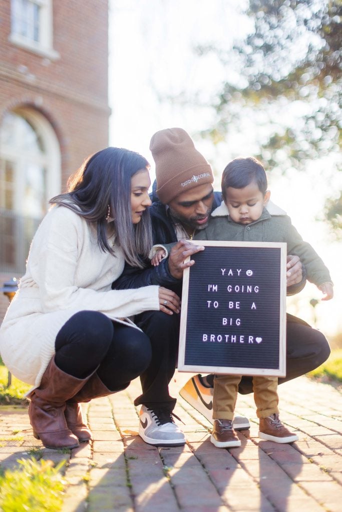 A couple kneels beside their child, who holds a sign reading "Yay, I'm going to be a big brother." The family is outdoors on a sunny day, capturing this precious moment in portraits at the picturesque Belair Mansion.