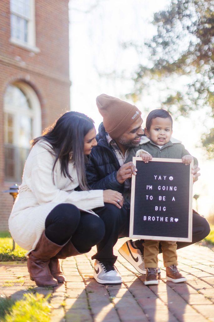 A couple crouching beside a young boy holding a sign that reads, "Yay! I'm going to be a big brother." The outdoor scene captures a beautiful family moment on a brick pathway in front of a building with trees in the background—perfect for cherished maternity portraits.