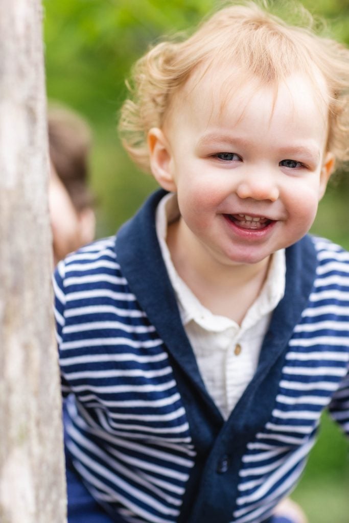 A young child with curly hair smiles while wearing a striped sweater. They are standing outdoors next to a tree, capturing a lovely family portrait at Brookside Gardens.