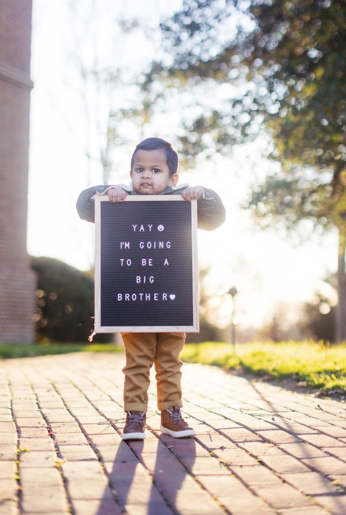 A young child holds a sign that reads, "Yay! I'm going to be a big brother," while standing on a brick path leading up to the historic Belair Mansion, with trees and greenery in the background.