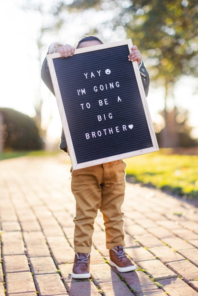 On a brick path, a child holds a letter board covering their face. The board reads, "Yay :) I'm going to be a big brother