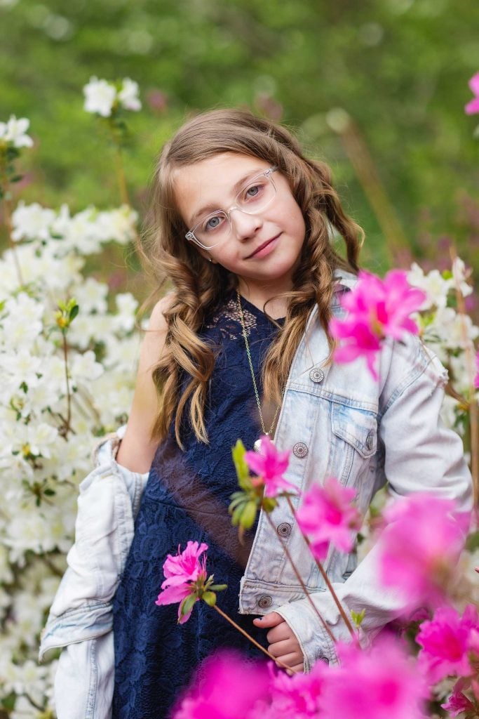 A young girl wearing glasses and a denim jacket over a blue dress stands among blooming flowers, looking at the camera, capturing a beautiful family portrait.