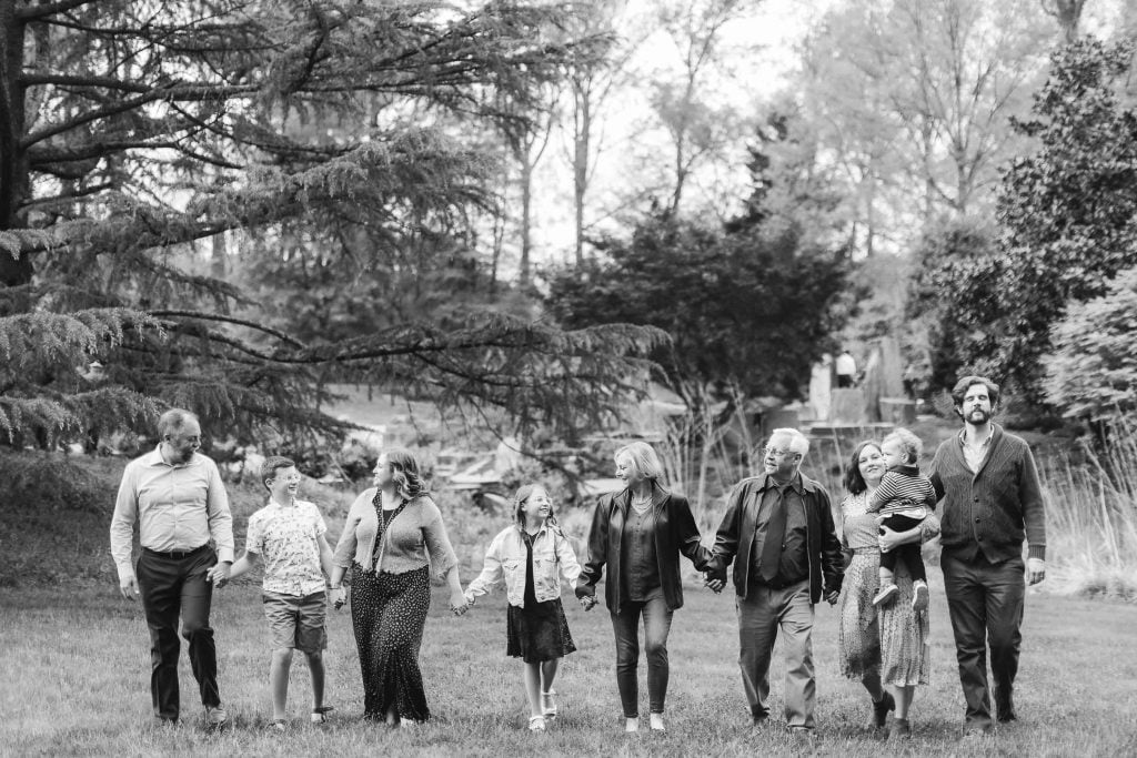 A blackandwhite photo captures an extended family of eight holding hands and walking through Brookside Gardens. Four adults walk on either side of four children, with trees visible in the background.