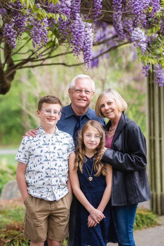 A smiling elderly couple stands under a wisteria tree with their two young grandchildren, a boy in a patterned shirt and khaki shorts, and a girl in a blue dress and glasses. This charming family portrait was captured at Brookside Gardens.