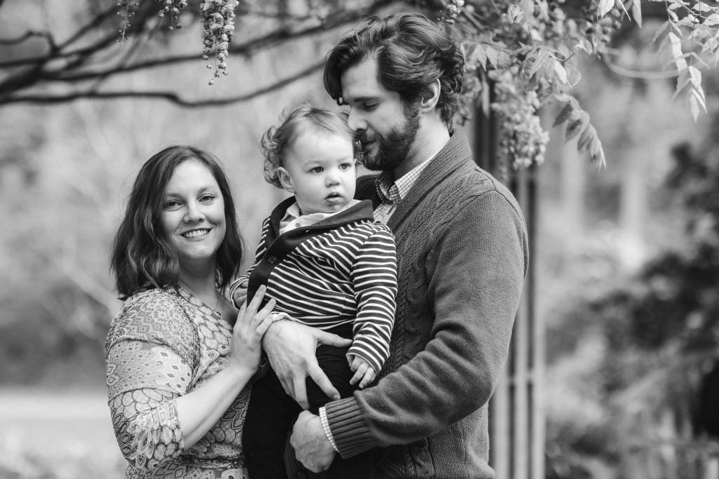 A family of three poses outdoors at Brookside Gardens. The mother smiles at the camera, while the father, holding their child, looks down. The child, dressed in stripes, looks off to the side. This timeless black and white portrait captures their loving bond amidst natural beauty.